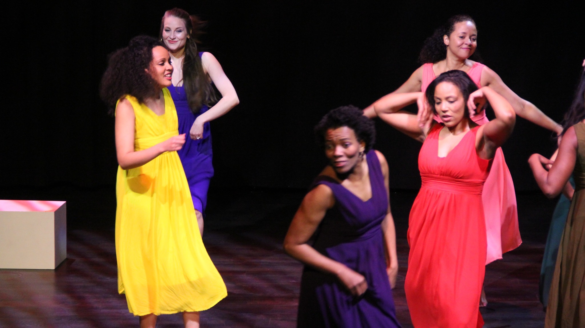 Women in yellow, coral, and purple moving across the stage in a bright dance image.