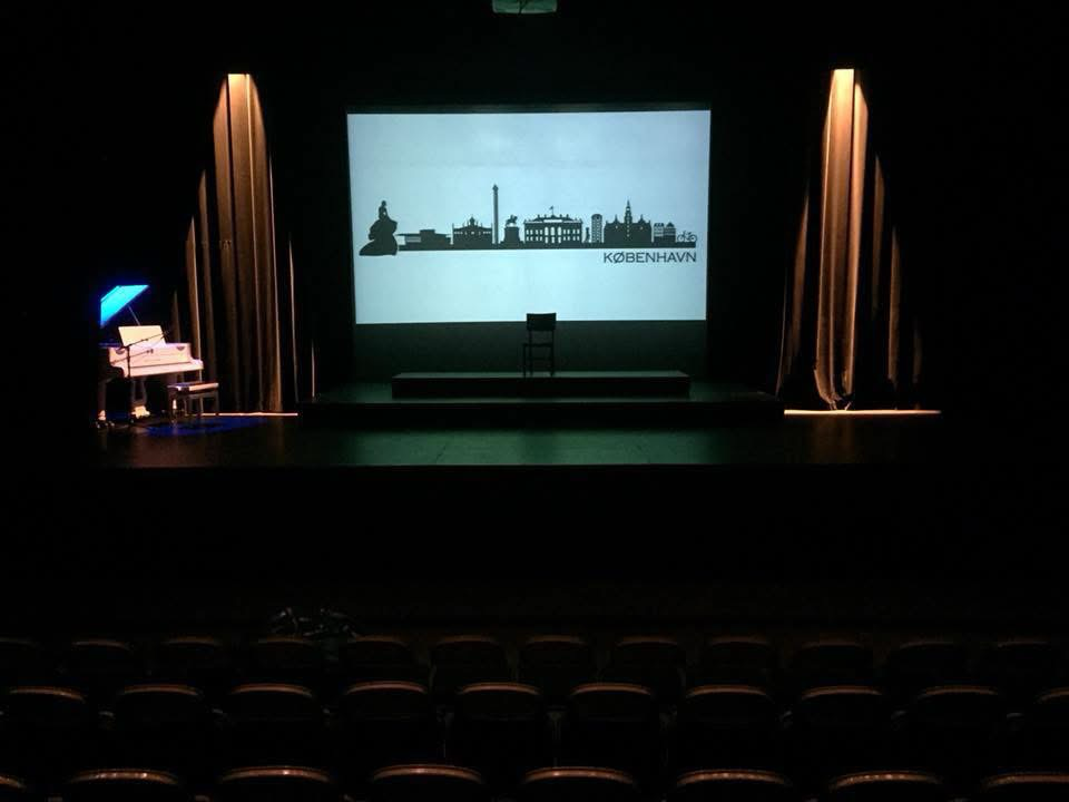 Wide stage photograph from The Merchant of Venice showing the Copenhagen skyline projection, piano, chair, and draped wings.